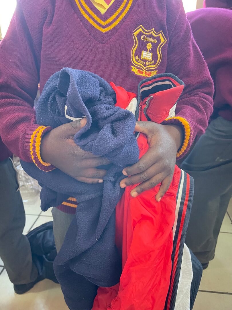 A Thuthuka Primary School learner in a new maroon ZS Uniforms jersey, holding the worn navy fleece and jacket worn before the handover, 30 July 2026